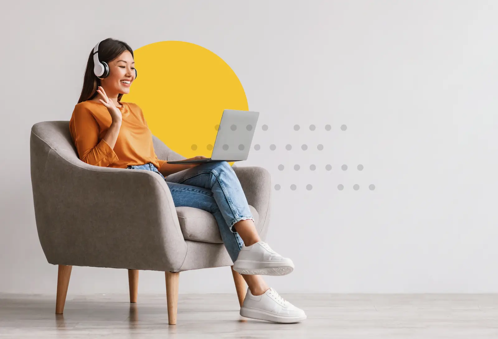 Woman in headphones waving during video call while using laptop in armchair with abstract yellow circle background.