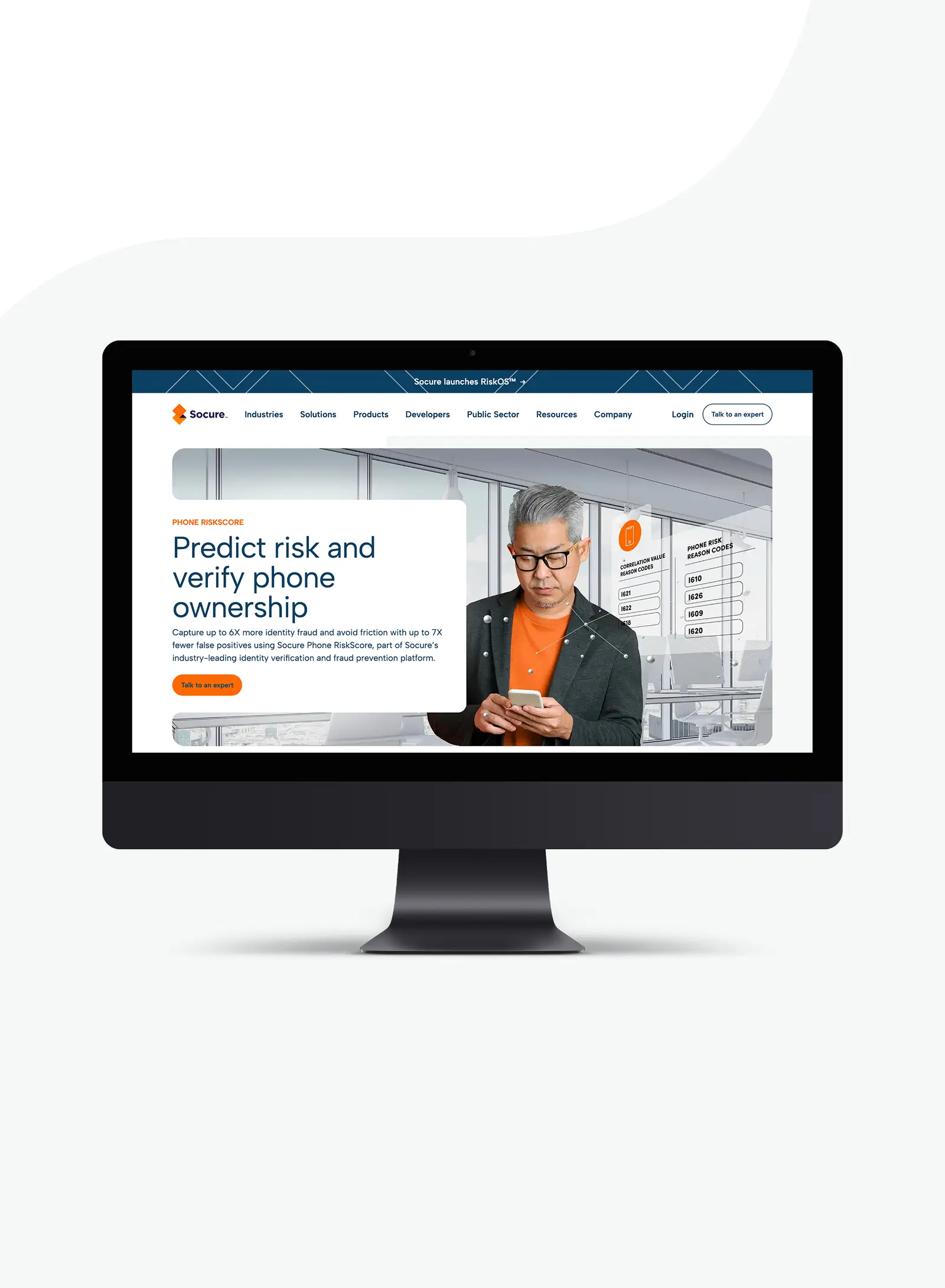 Desktop monitor displaying a phone risk verification platform, with a man in glasses looking at his smartphone beside on-screen charts about predicting risk and verifying phone ownership.