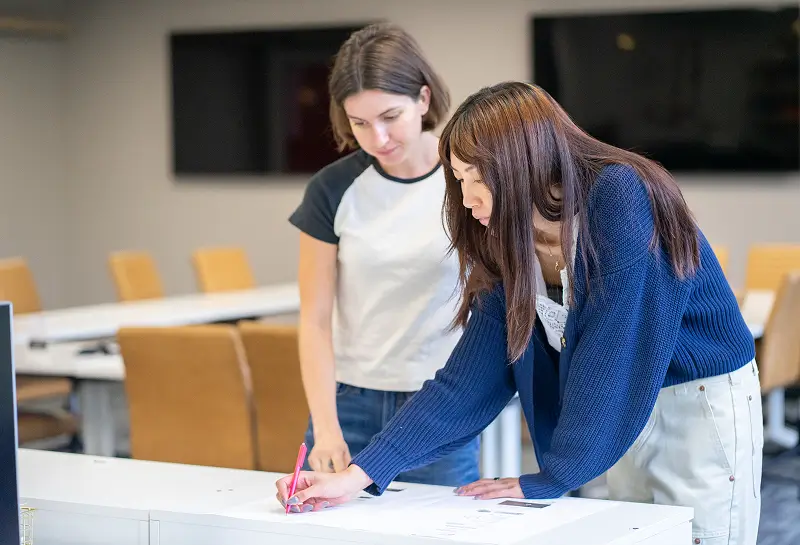 Two students working together at a desk in a classroom, with one writing on paper while the other observes.
