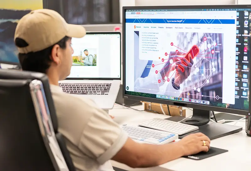Person in a cap working at a desk with a laptop and a large monitor displaying a digital network interface.