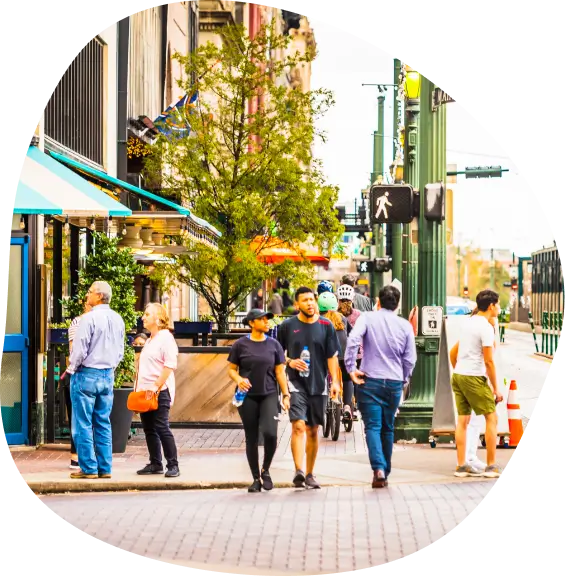 People walking and standing along a lively city street lined with shops, outdoor seating, and trees on a bright day.