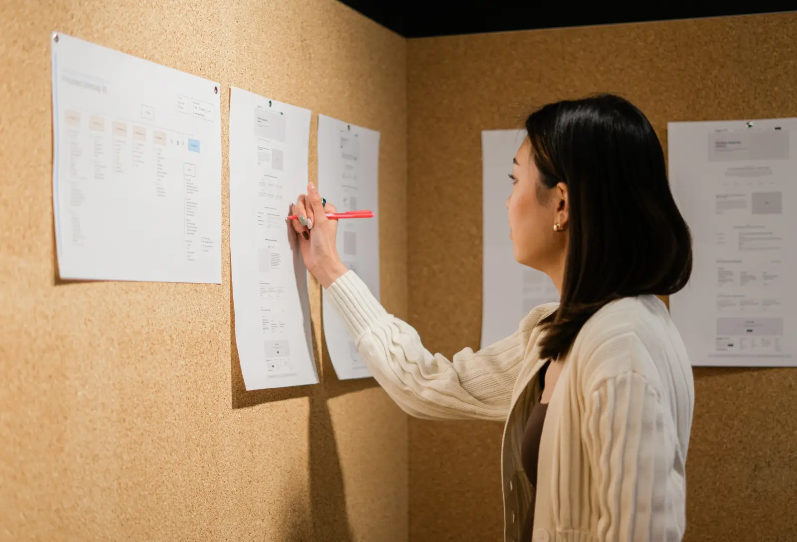 Woman reviewing printed wireframes on a corkboard and marking notes with a pen