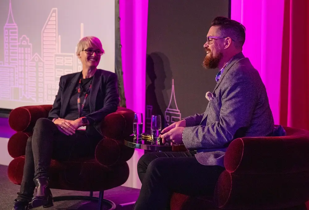 Two speakers in armchairs engaged in a panel discussion on a stage with purple lighting and a city-themed backdrop.