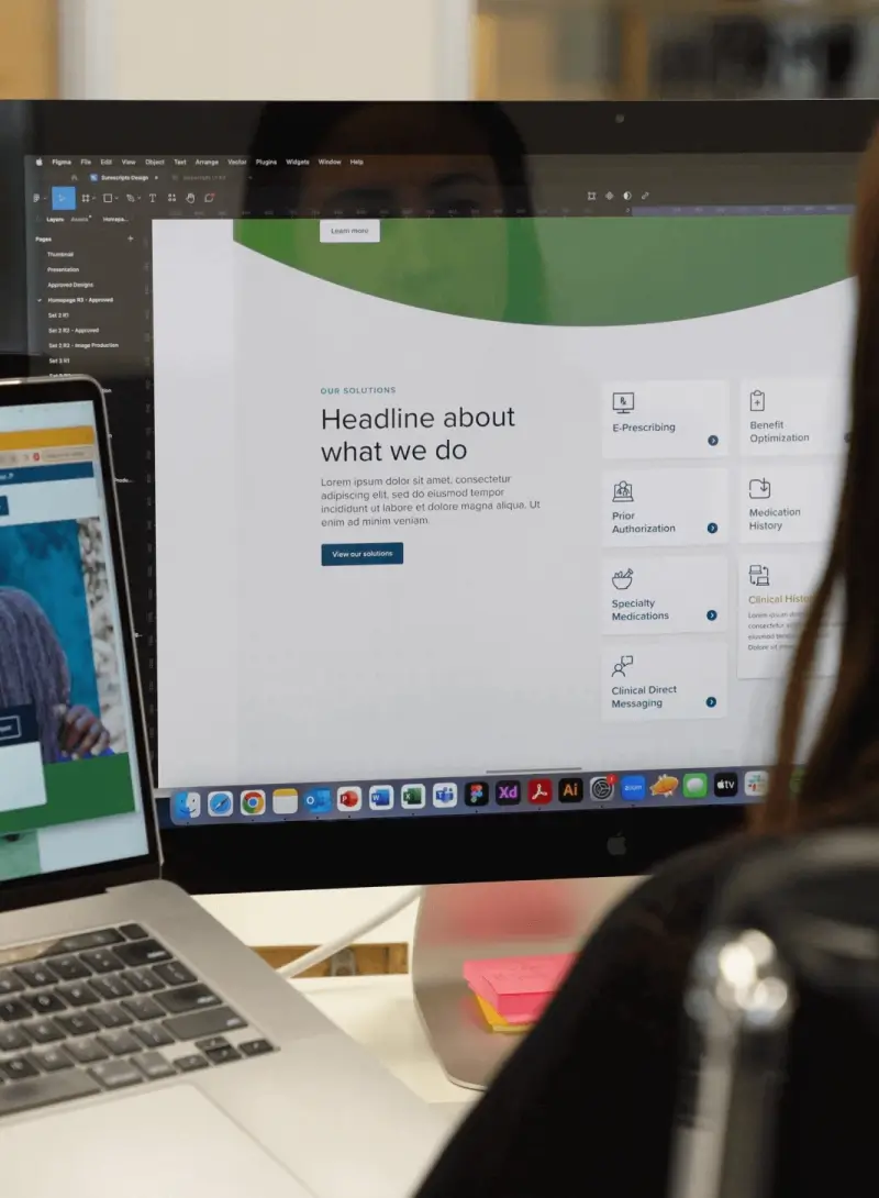 Designer working at a desktop computer with a website layout open, alongside a laptop, in a modern workspace.