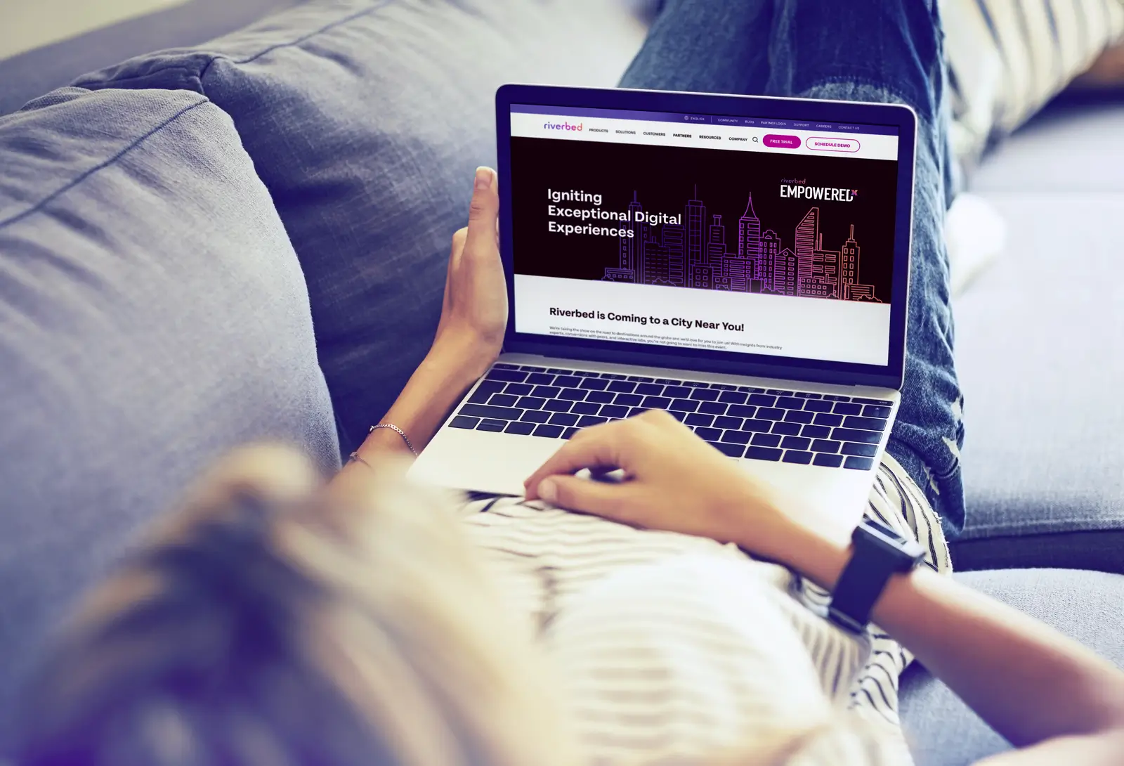 Person using a laptop on a couch displaying the Riverbed Empowered event landing page with digital experience branding.